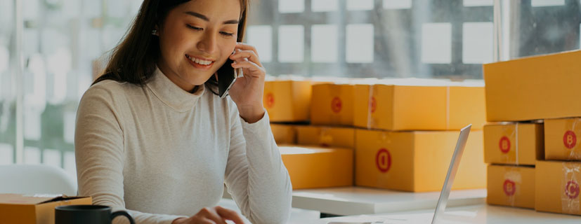 Imagem de uma mulher falando no celular, sentada em frente a uma mesa com vários papéis