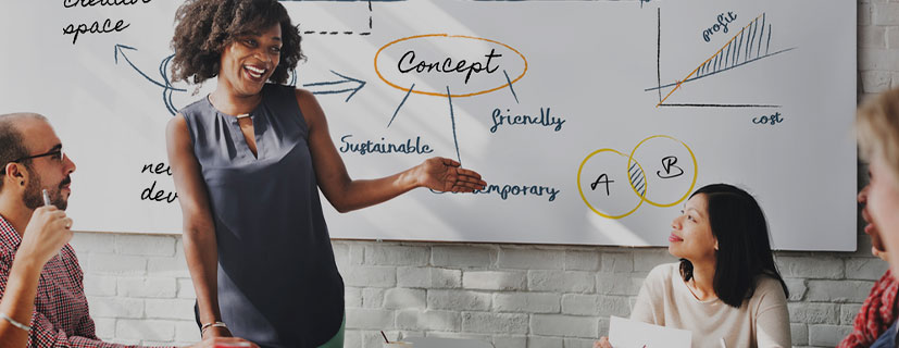 Imagem de uma mulher apontando para uma lousa cheia de estratégias com uma equipe assistindo