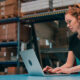 mulher digitando no notebook que está em cima de uma mesa dentro de um estoque