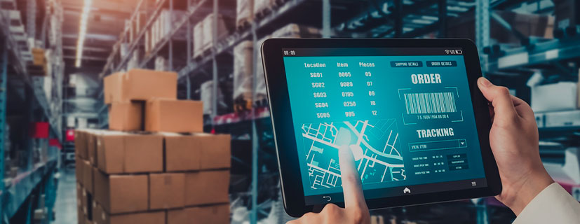 Imagem de mãos segurando um tablet e apontando na tela, dentro um galpão cheio de caixas