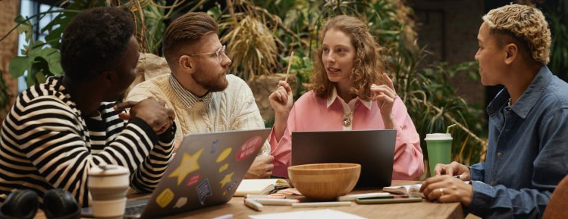 Pessoas conversando sentados em volta de uma mesa cheia de notebooks e itens de trabalho de escritório