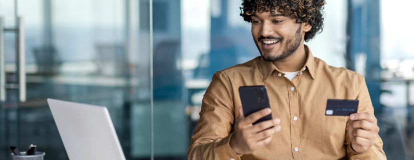 Homem usando o notebook com um celular e um cartão em mãos.